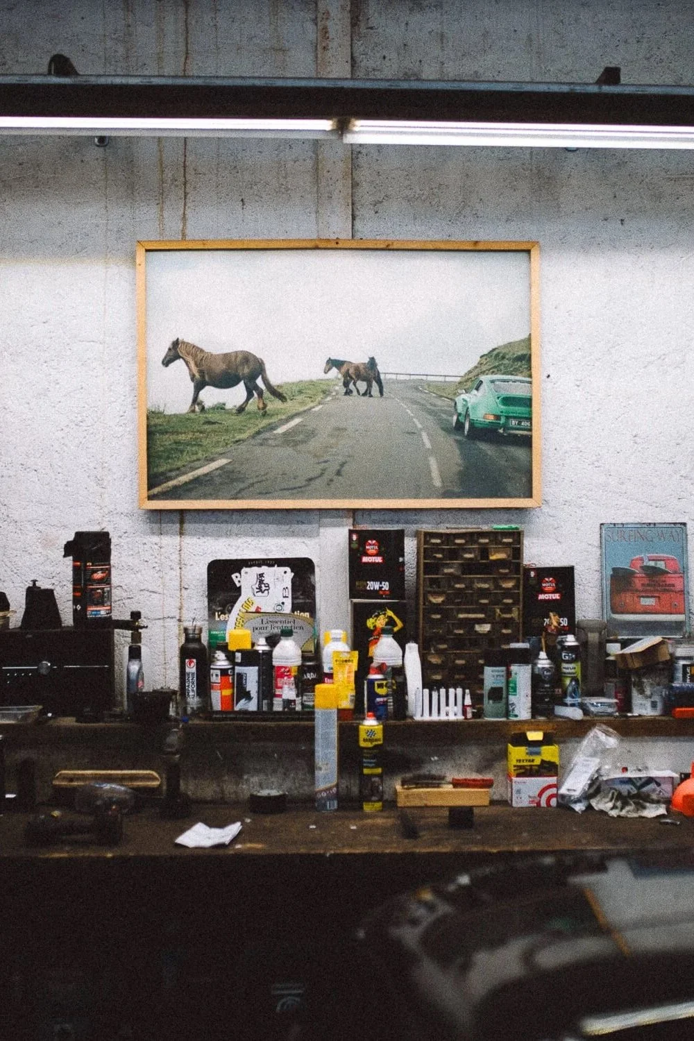 A cluttered workshop with tools, containers, and a framed photograph of three horses and a vintage car on a winding road.