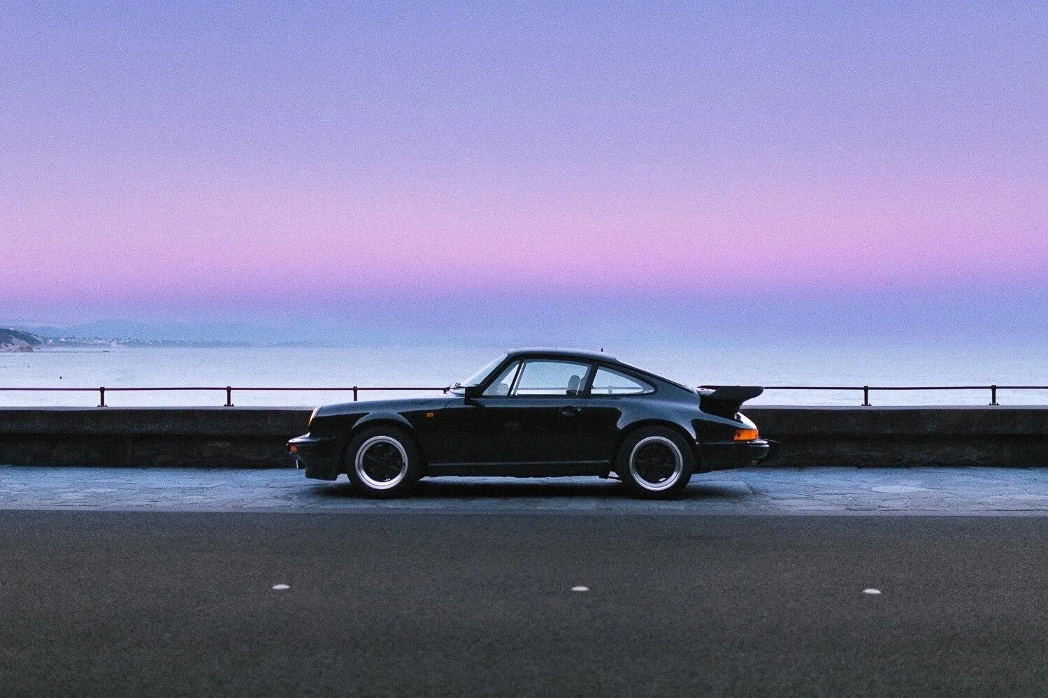 A black vintage Porsche 911 parked near a seaside promenade at dusk with pastel purple and pink sky in the background.