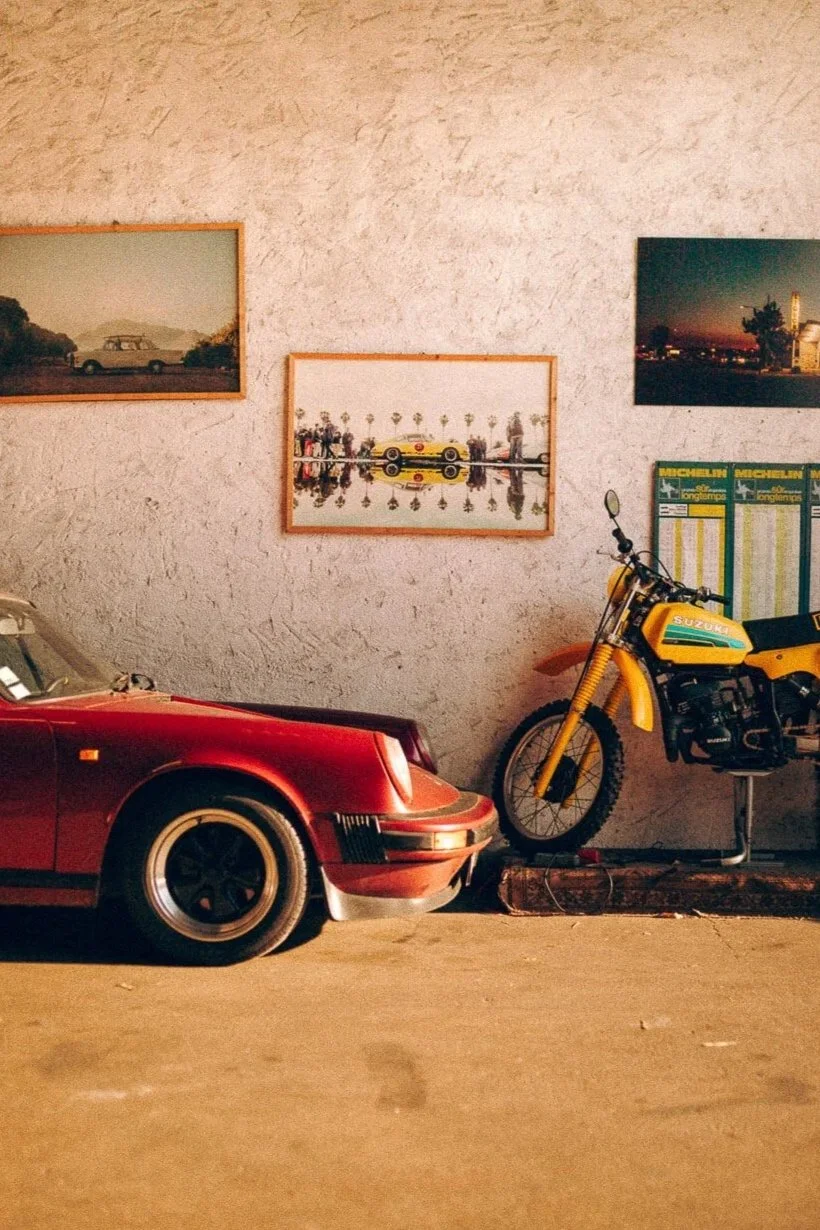 A red vintage car and a yellow Suzuka motorcycle on display against a textured wall decorated with framed photographs and a Michelin poster.