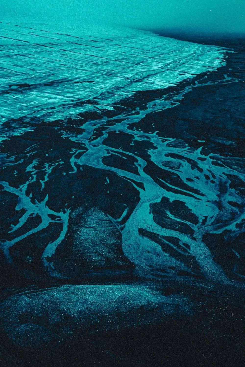 Vue aérienne artistique de motifs de rivières sinueuses rencontrant une mer ou un océan, avec des teintes bleutées et un contraste élevé.