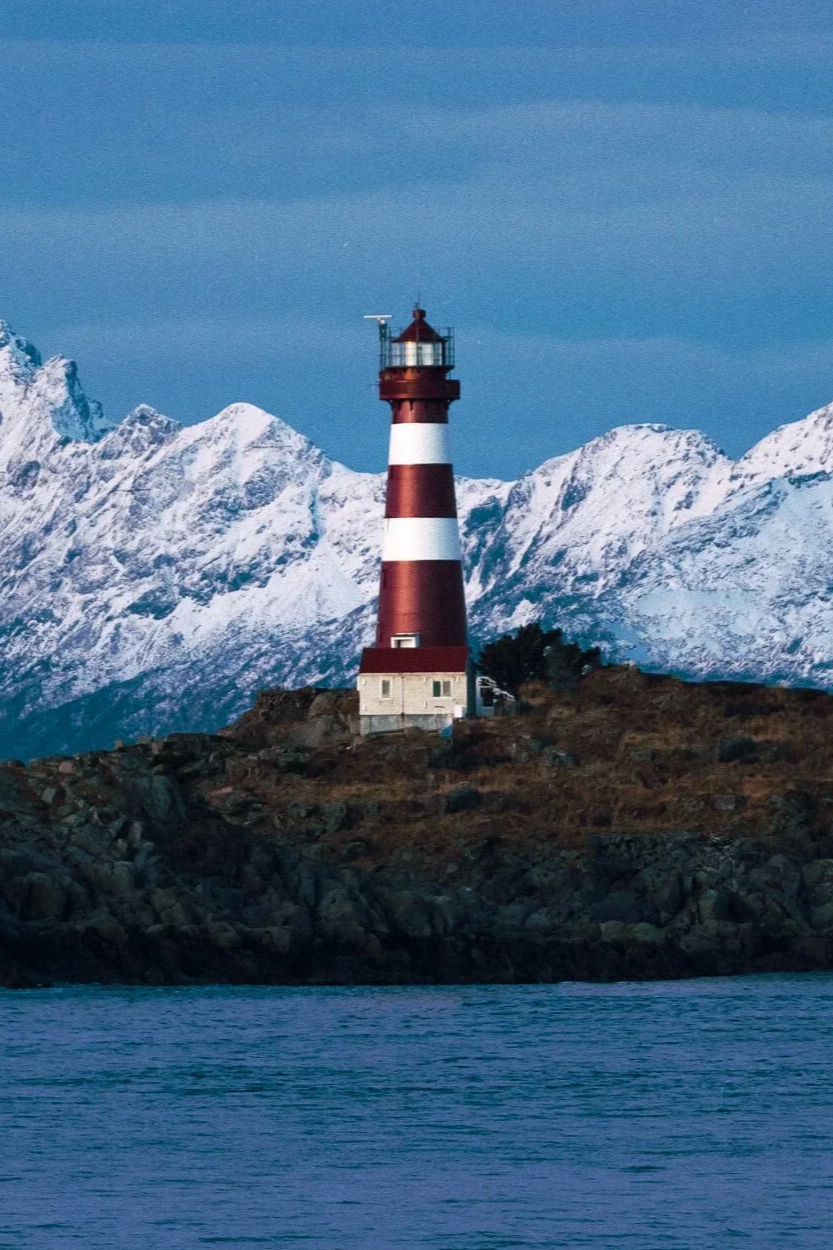 Phare rouge et blanc sur une île rocheuse, avec des montagnes enneigées en arrière-plan et l'eau au premier plan.