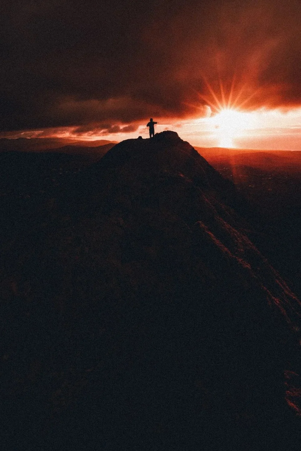 Personne debout sur un sommet, silhouette contre un coucher de soleil dramatique avec des nuages sombres et rayons de lumière éclatants.