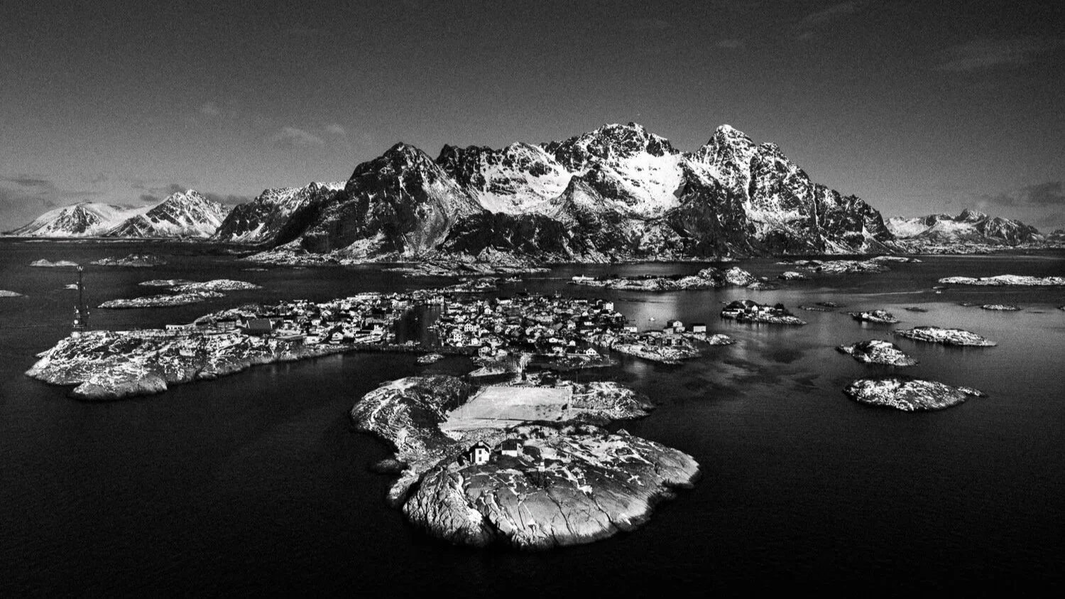 Paysage montagneux enneigé avec petites maisons sur des îles entourées d'eau, vue d'en haut en noir et blanc.