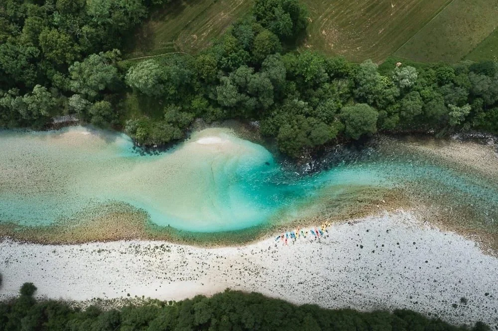 Aerial view of a river with turquoise water flowing through a forested area, with a sandy beach and people gathering near the shore.