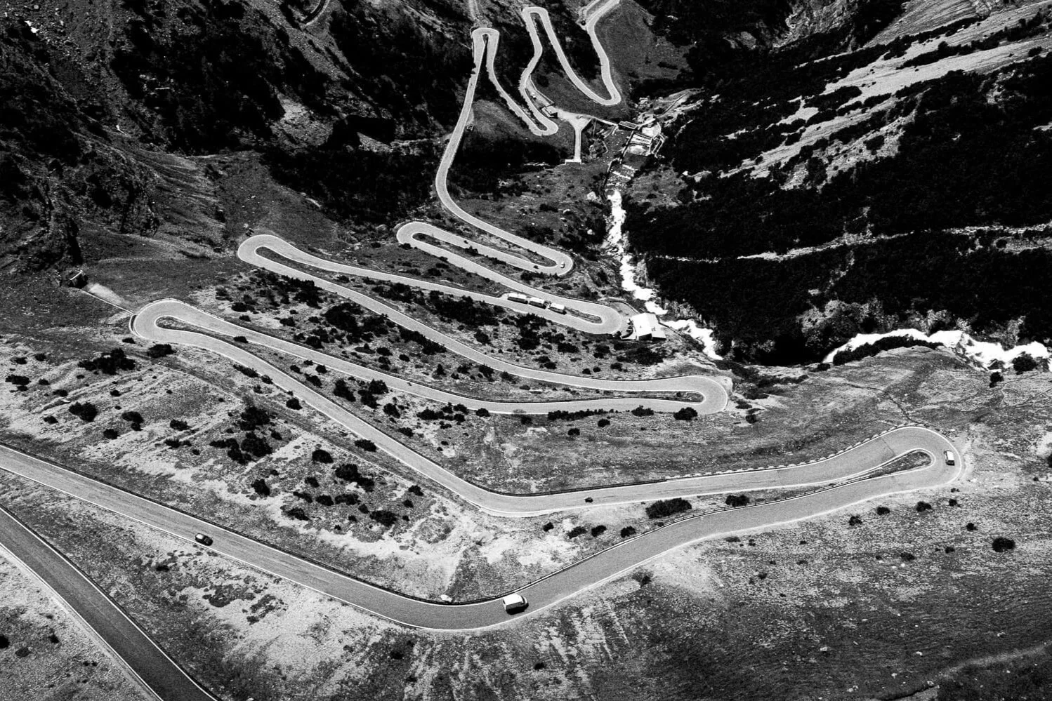 Black and white aerial photograph of a winding mountain road with multiple switchbacks. Several vehicles are driving along the road, and a river runs through the valley below.