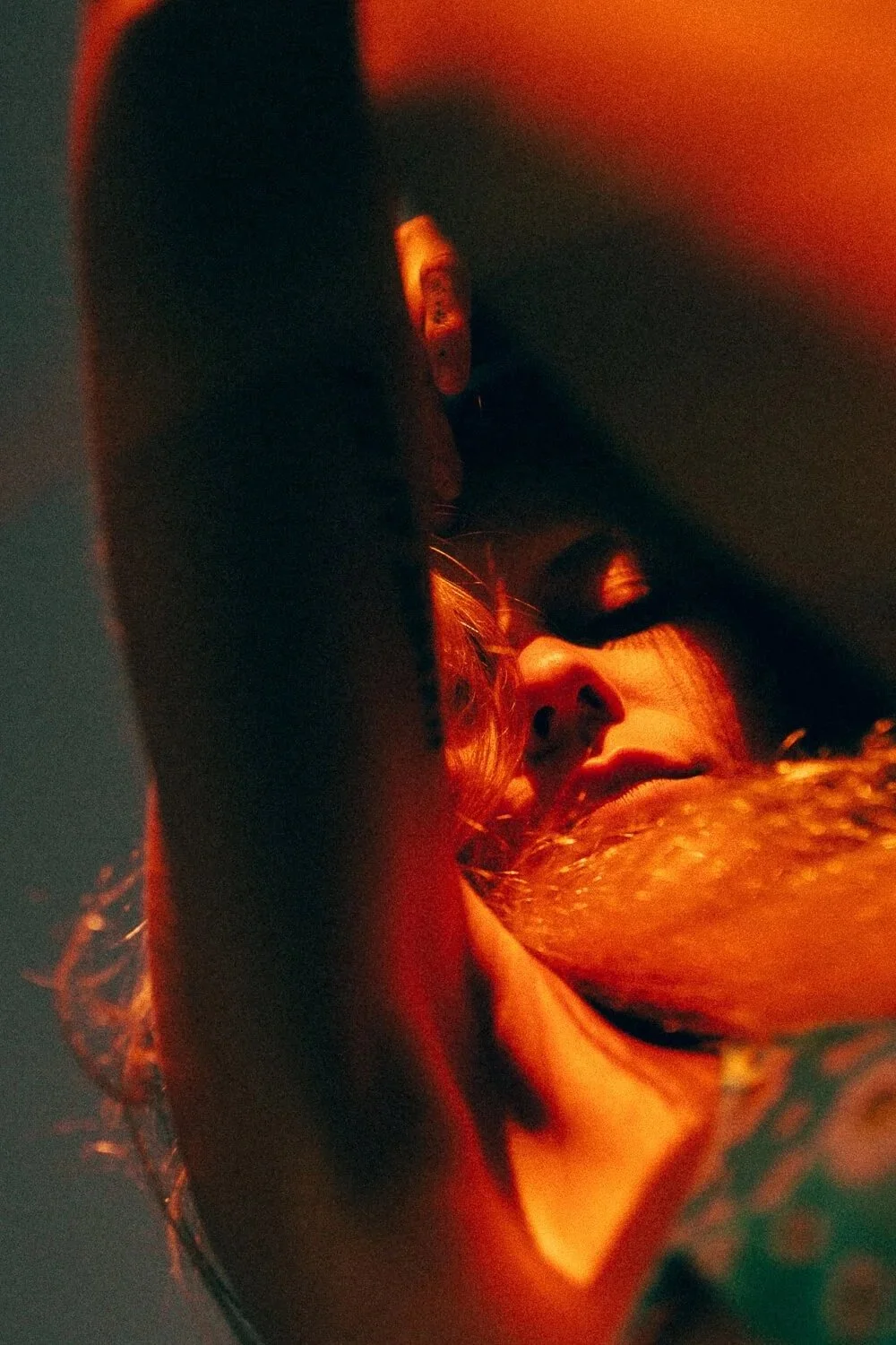 A close-up of a person's face, with long hair, in low lighting that gives an orange-red hue, partially obscured by their arm.