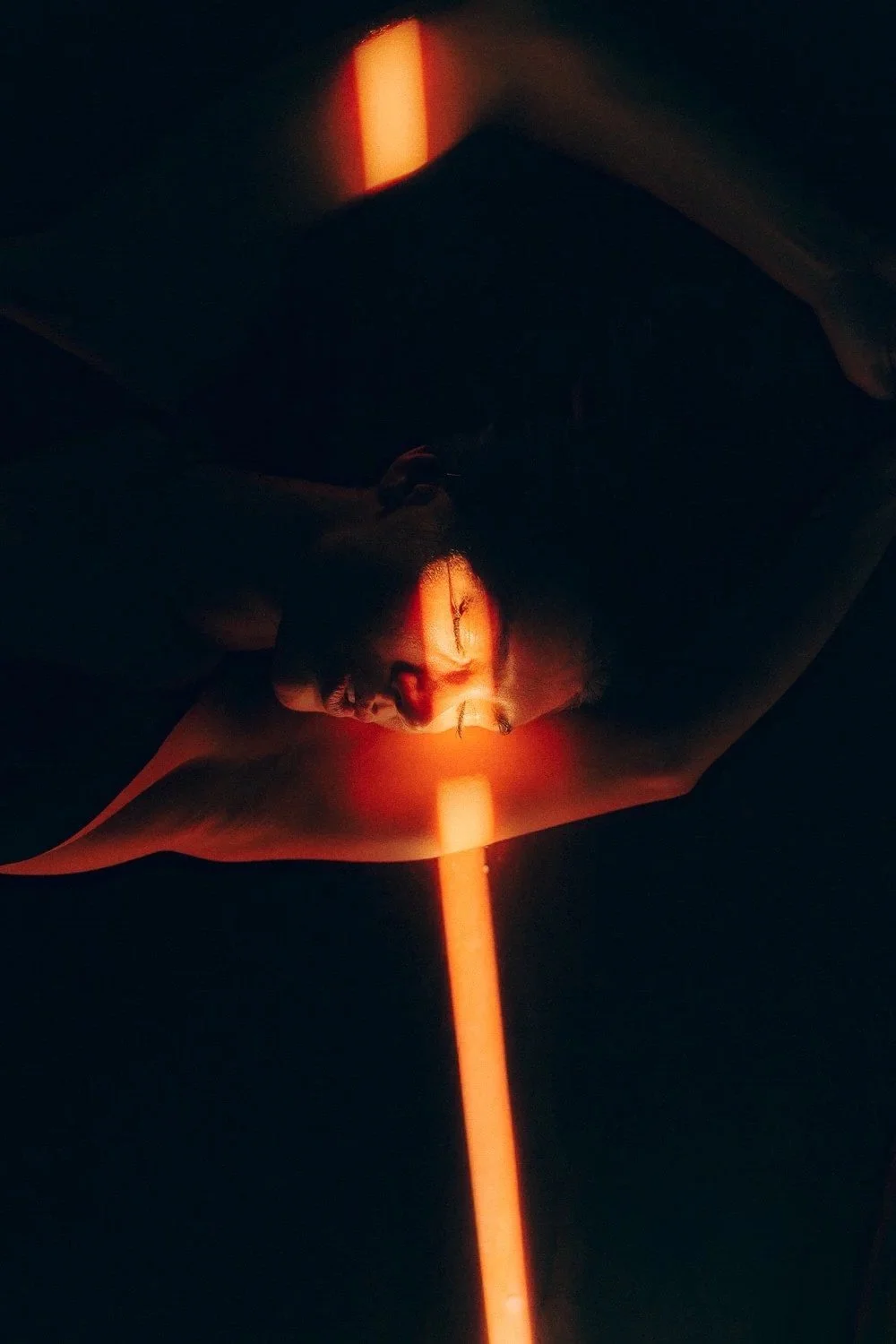 Person's face reflected on a mirror with a bright orange laser beam running vertically through the image, in a dark room.