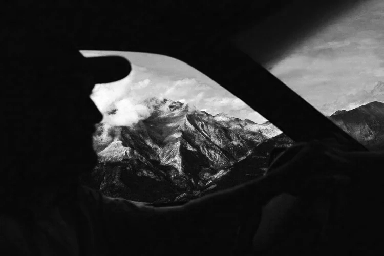 Silhouette d'une personne au volant d'une voiture, vue à travers le pare-brise vers un paysage montagneux au coucher du soleil.