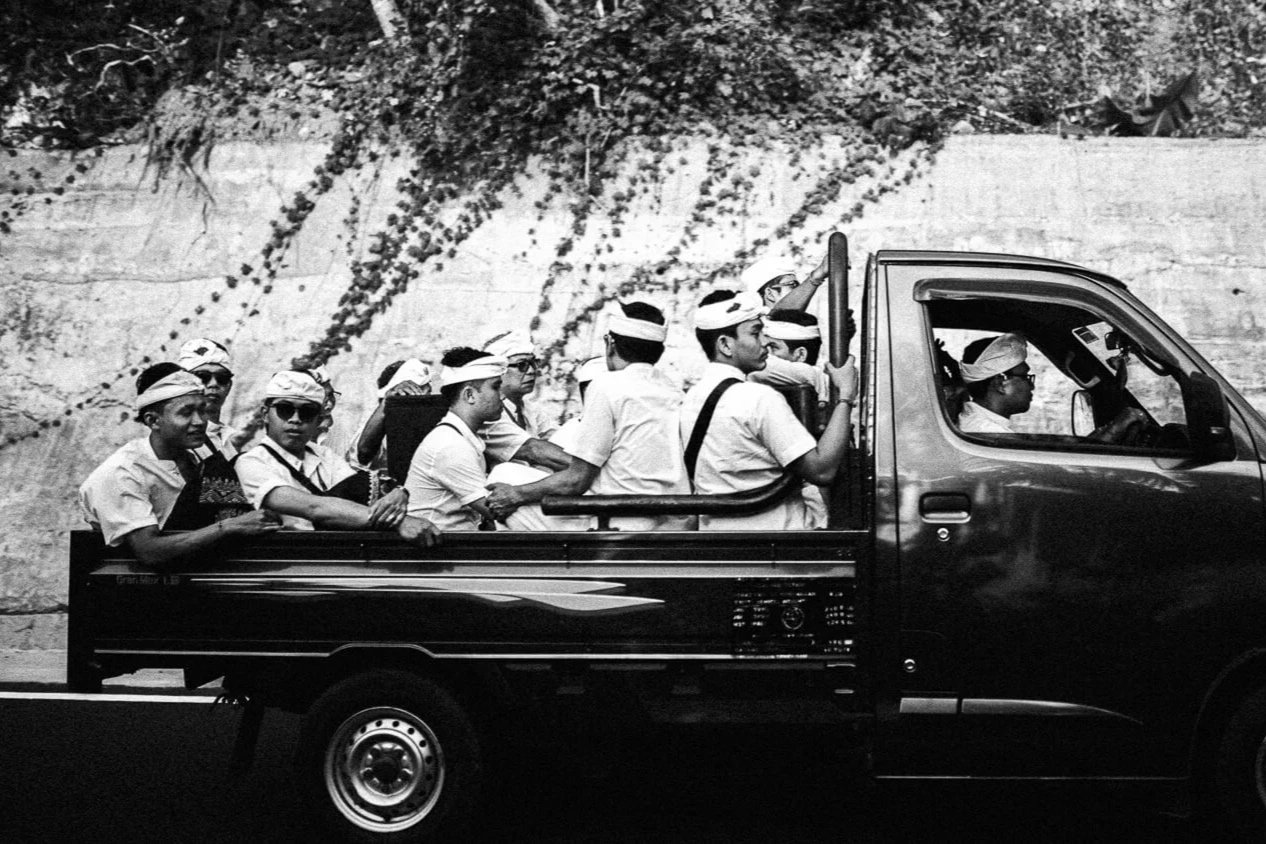 Groupe de personnes assises dans la caisse ouverte d'un petit camion, portant des vêtements traditionnels et des bandeaux, sur un fond de roche et de végétation.