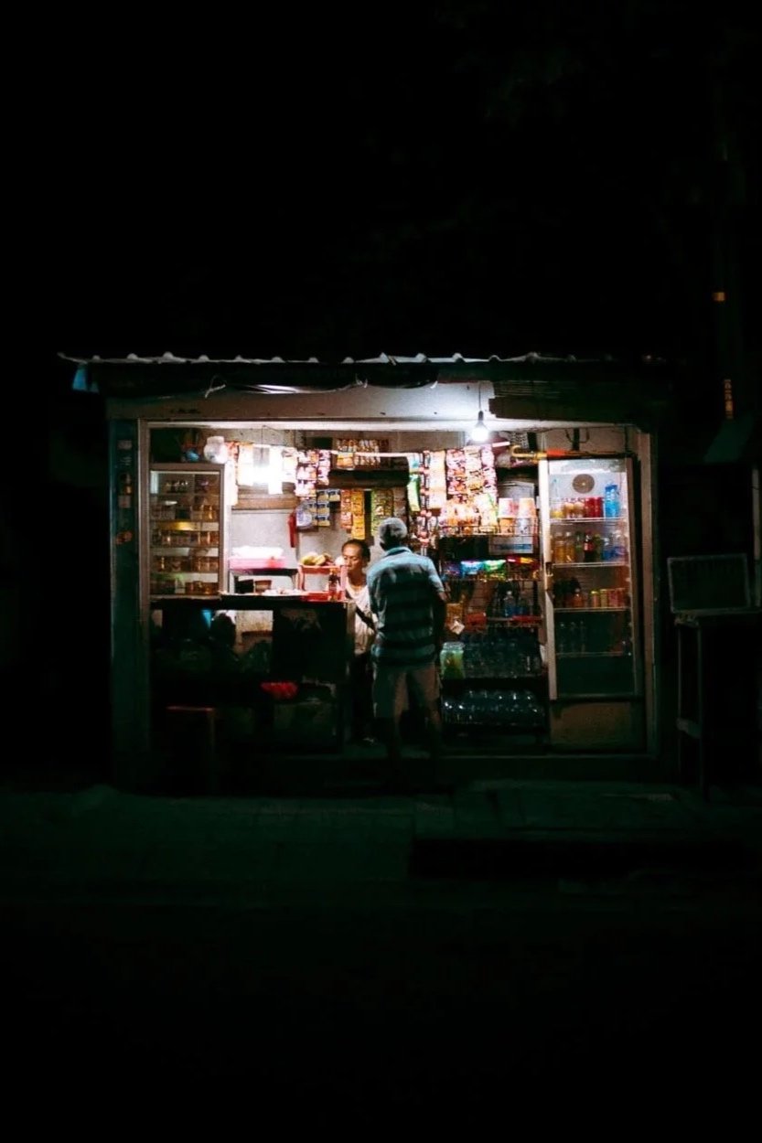Une petite boutique éclairée la nuit avec deux personnes à l'intérieur, vendant divers produits comme des snacks, boissons et autres articles de commerce.