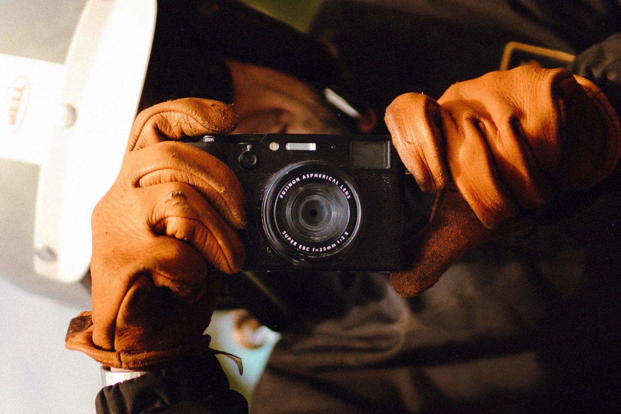 Person wearing tan gloves taking a mirror selfie with a black Fujifilm camera indoors, with part of a white wall and a gray surface visible in the background.