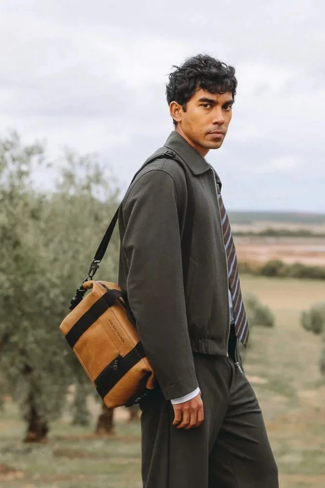 A man in a dark jacket and tie walking through an olive grove with a tan shoulder bag.