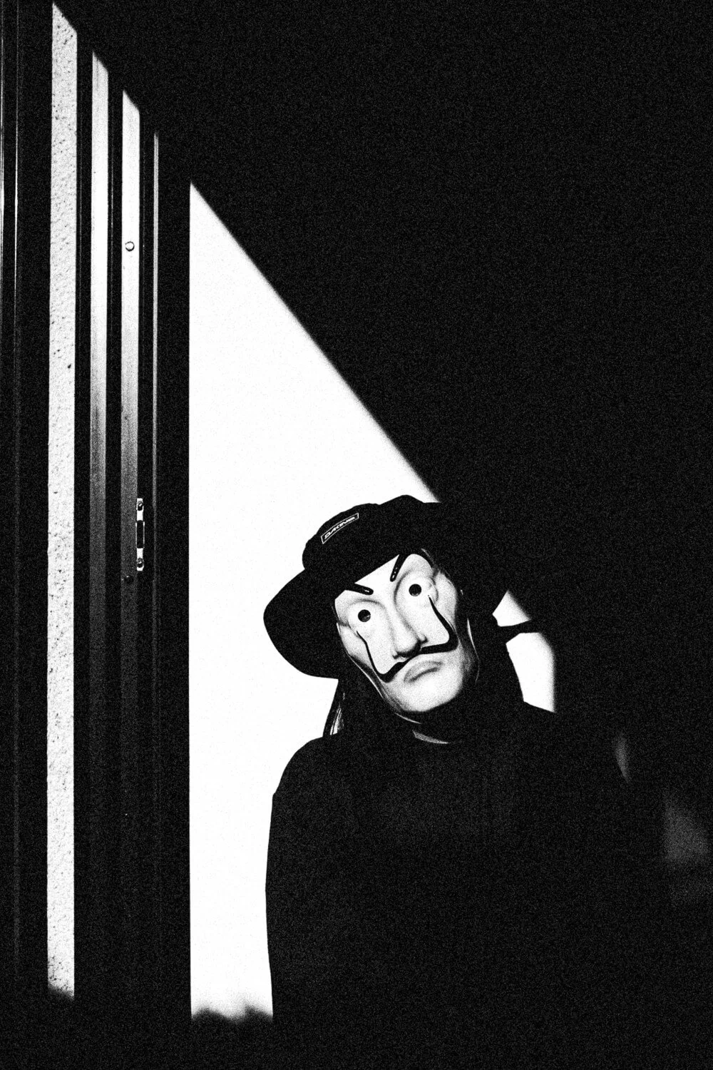 Person wearing a Guy Fawkes mask and a hat, standing in a doorway with lighting creating a shadow on the wall.