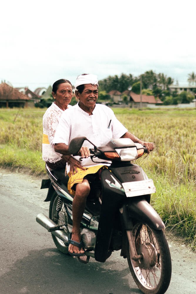 An elderly man and woman riding a motorcycle together through a rural area with houses and greenery in the background.