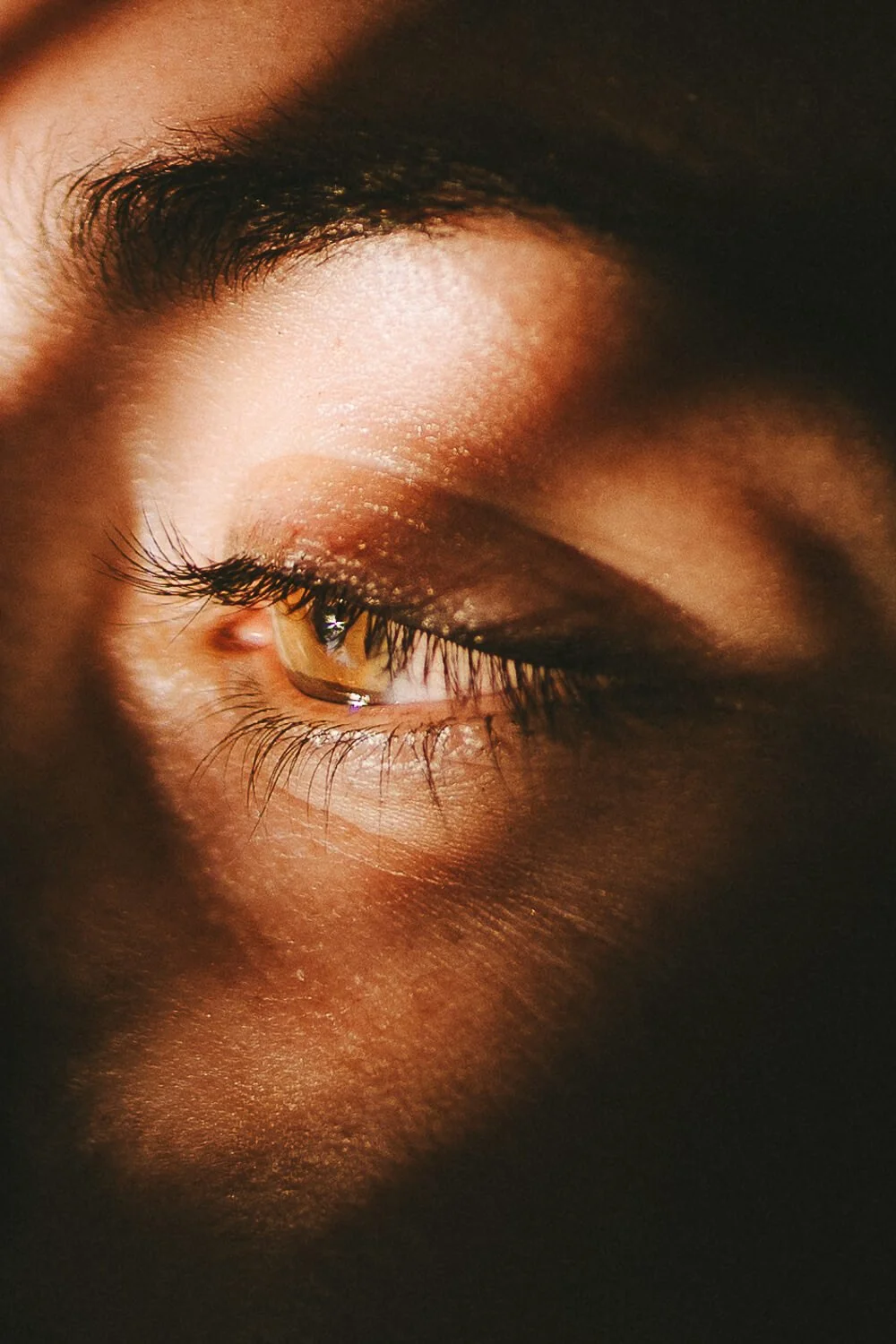 Close-up of a person's face with one eye closed, showing detailed eyelashes, eyelid, and eyebrow, with warm lighting highlighting the skin's texture.