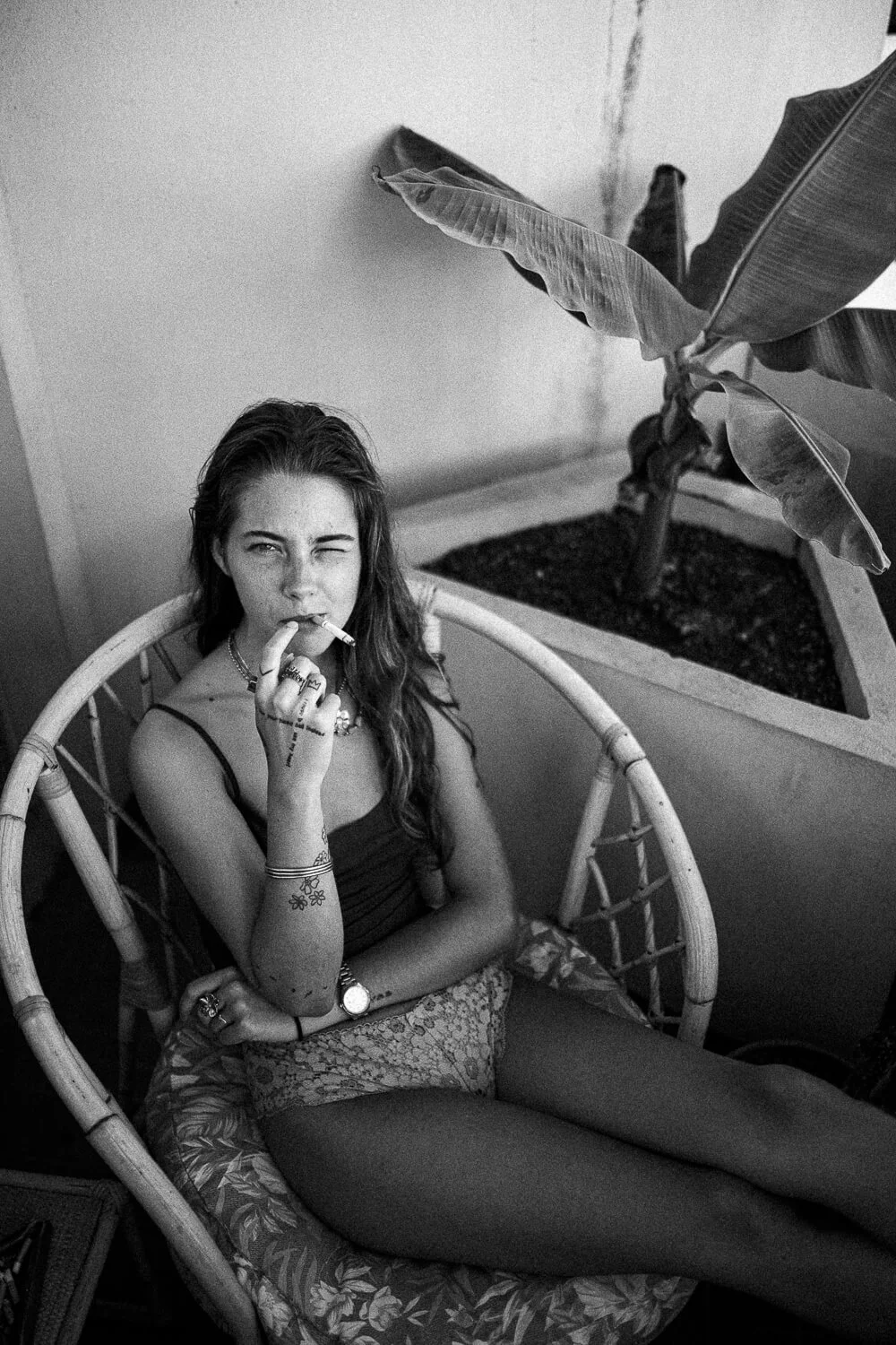 A young woman sitting on a wicker chair, holding a cigarette, with one eye closed. She has tattoos on her arm, wears rings, a watch, and a necklace, and is dressed in a tank top and shorts. There is a large potted plant nearby.