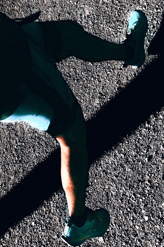 Close-up of a person's legs and feet, wearing athletic shoes, on a gravel surface with long shadows.