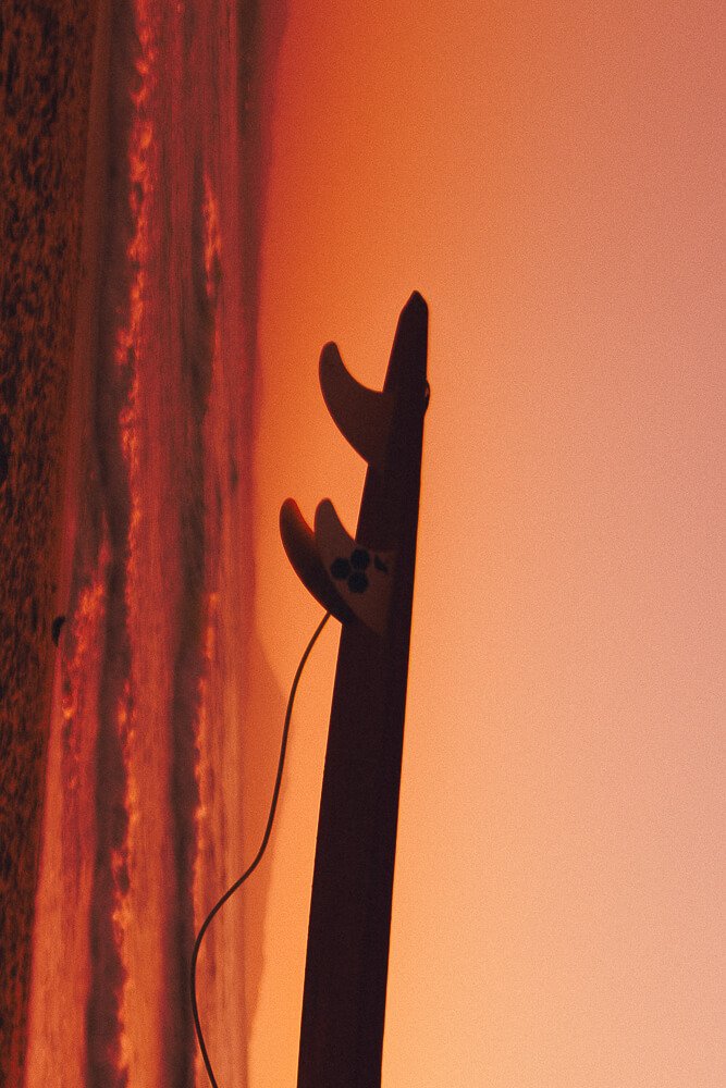 Silhouette of a surfboard with fins, standing upright on the beach during sunset