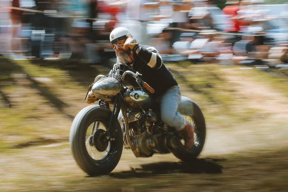 Motorcyclist in vintage gear riding on a dirt track with blurred spectators in the background.