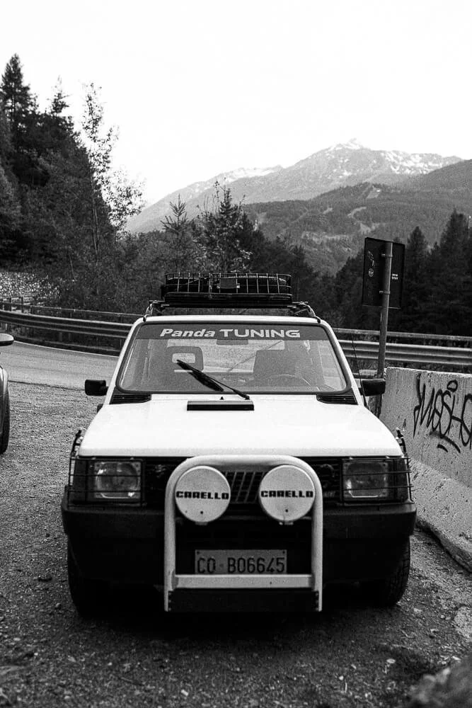 Black and white photo of a car with a roof rack and front-mounted rally lights, parked on a mountain road with a mountainous landscape in the background.