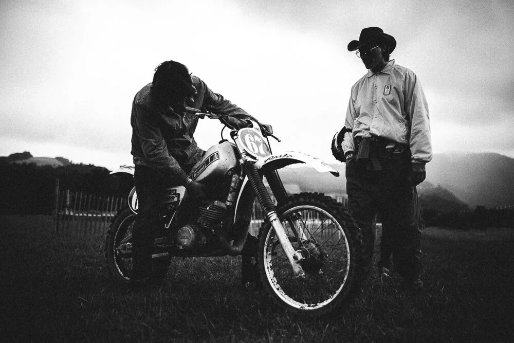 Two people, one on a dirt bike and one standing next to it, are outdoors on a grassy field with mountains in the background. The photo is in black and white.
