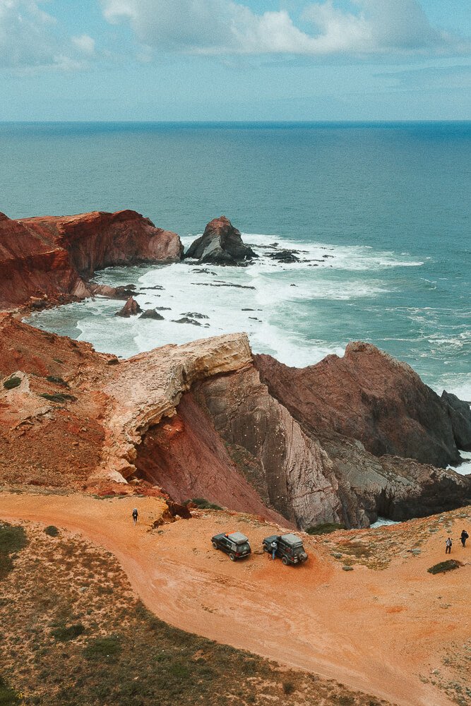 Cliffside view of rugged coastline with red and brown rock formations, two off-road vehicles parked on dirt trail, and people walking along the edge, overlooking the ocean with waves crashing against rocks.