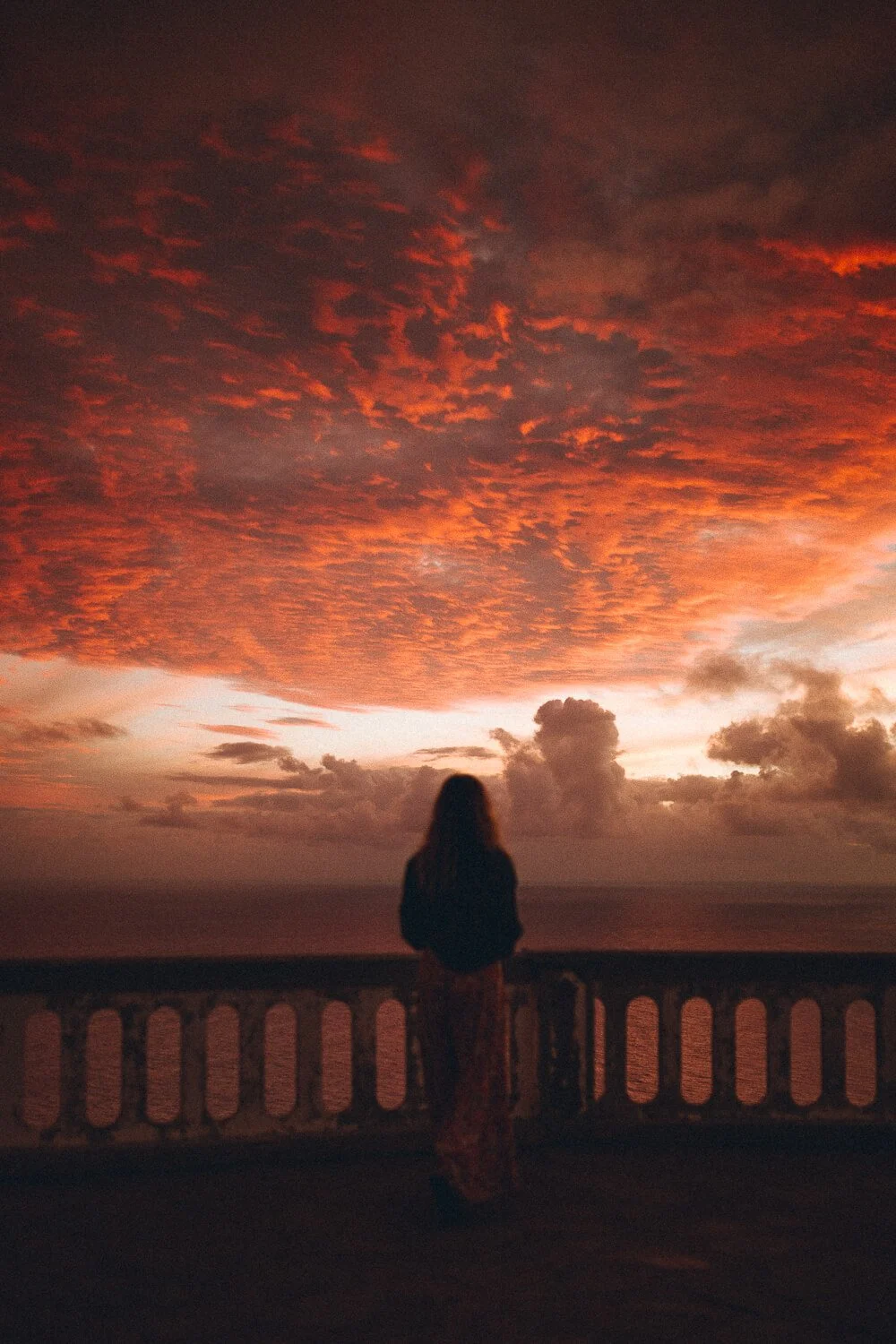 A person standing on a balcony overlooking the ocean at sunset with a vibrant orange and red cloud-filled sky.