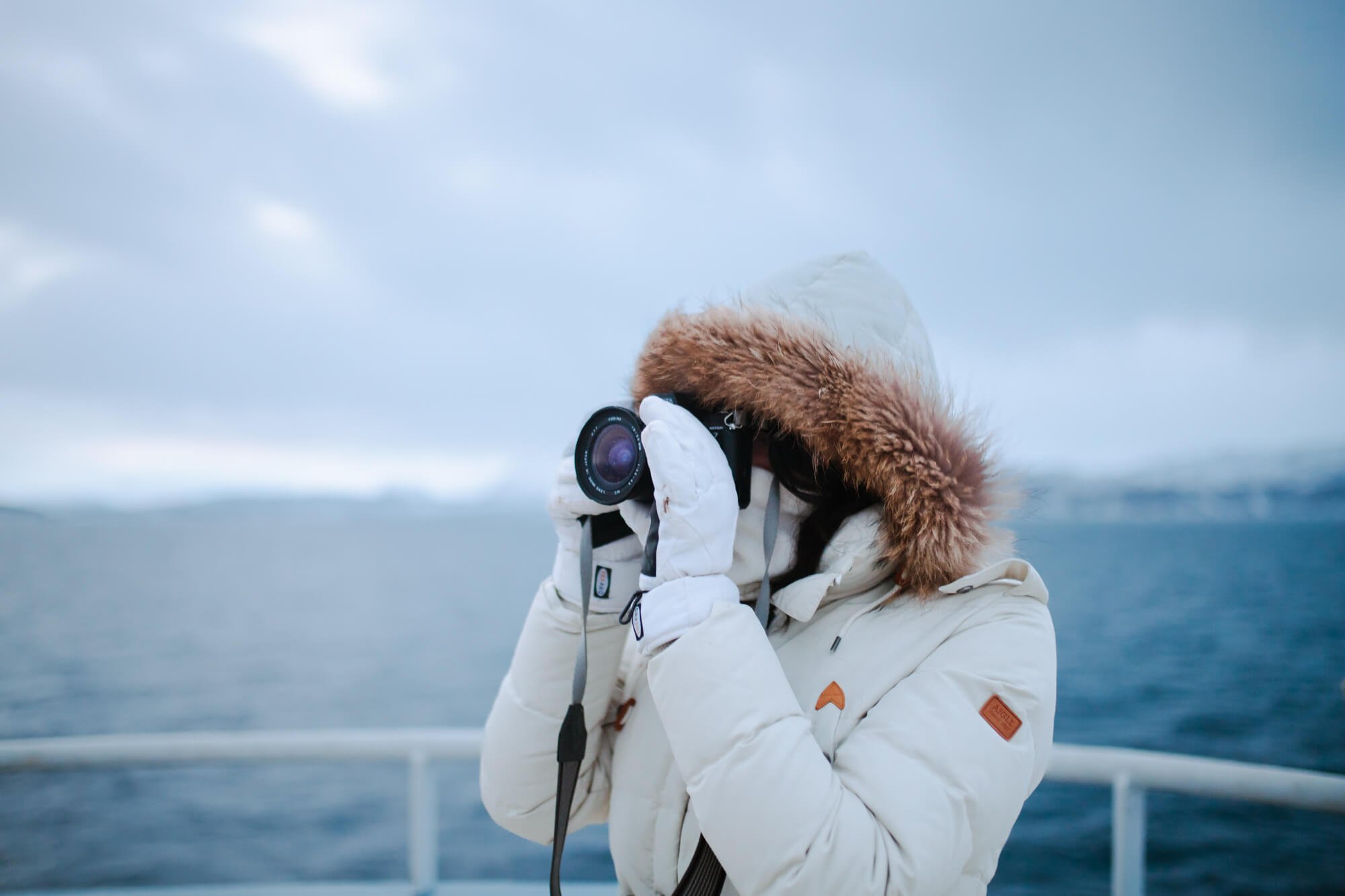 Une personne en manteau d'hiver blanc avec capuche prend des photos avec un appareil photo près d'une étendue d'eau glacée.
