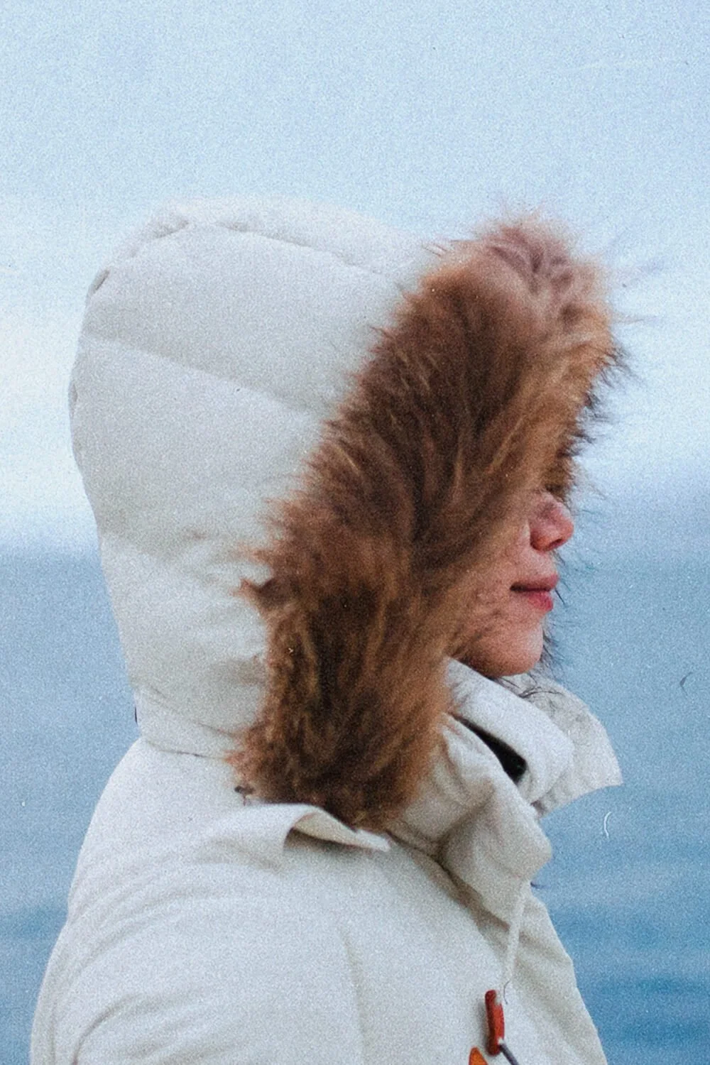 Femme portant une parka blanche avec une capuche en fourrure brune, faisant face au vent ou au froid.