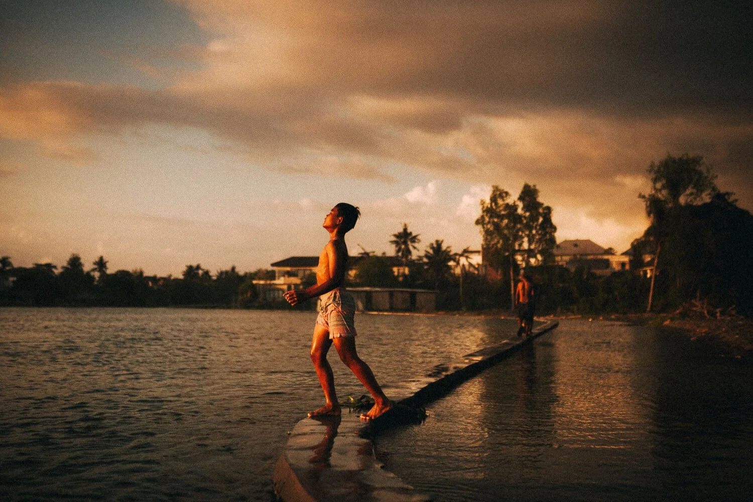 Un garçon marche sur une planche au coucher du soleil au bord de l'eau, avec des maisons et des arbres en arrière-plan.