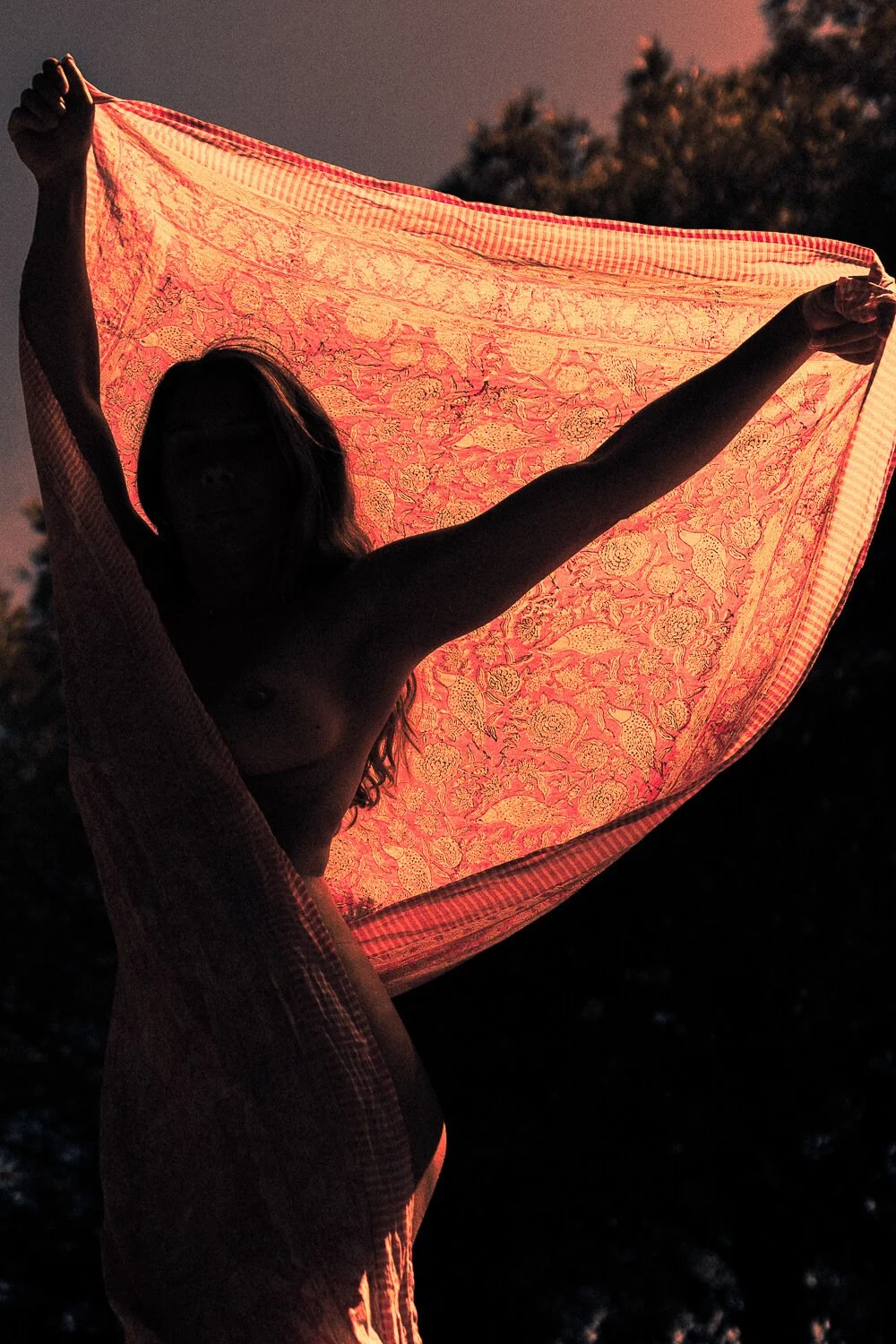 A person holding a colorful, patterned cloth or blanket against the sunset sky, with trees in the background.