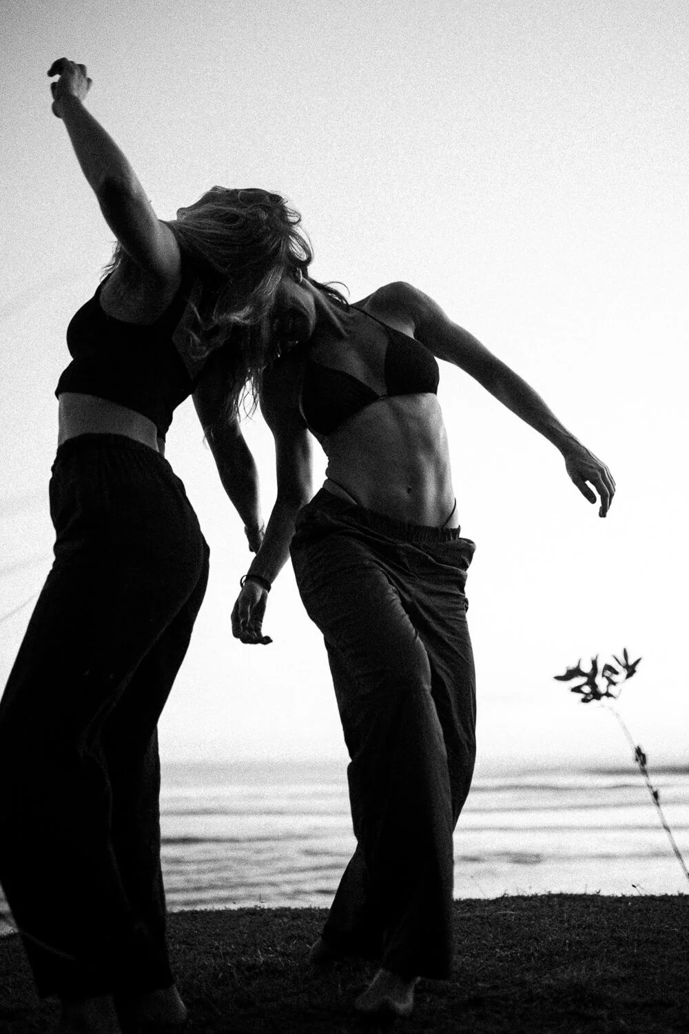 Two women dancing on the beach at sunset, black and white photograph, casual clothing, one in a sports bra and pants, showing movement and expression.