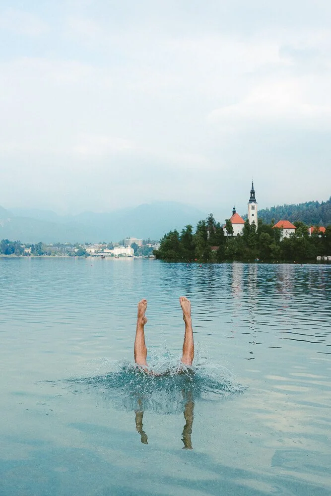 Person diving into a lake with legs and arms visible above the water, overlooking a small island with a church and trees, and mountains in the background.