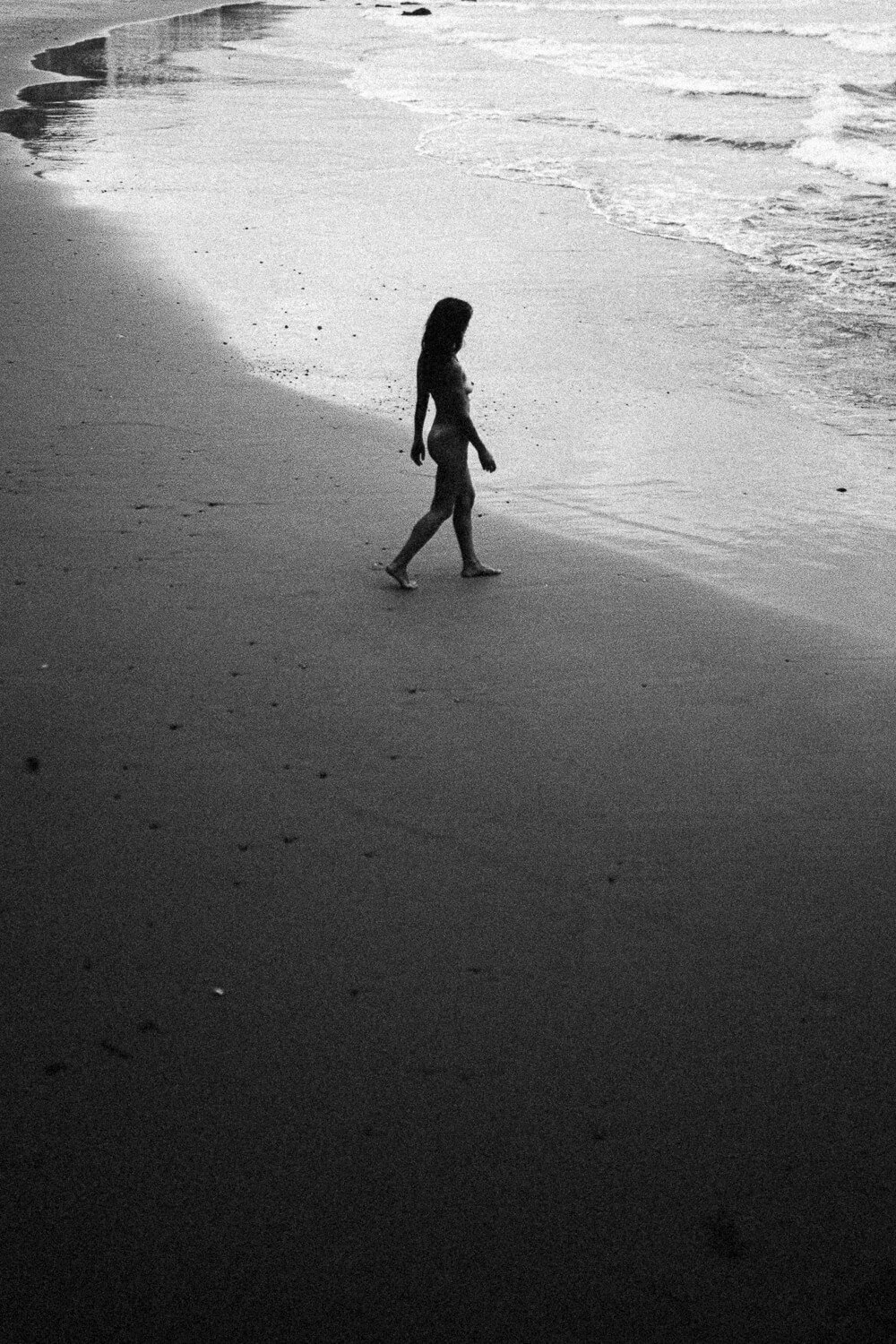 A silhouette of a person walking along the shoreline of a beach, with waves in the background during the day.