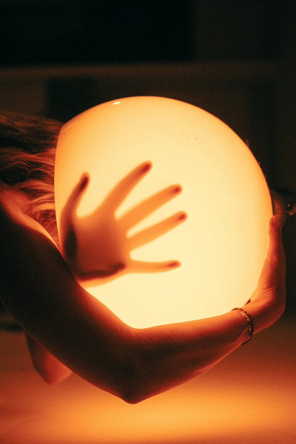 Person's hand and arm holding a glowing round lamp with fingers resting on its surface in a dimly lit room.