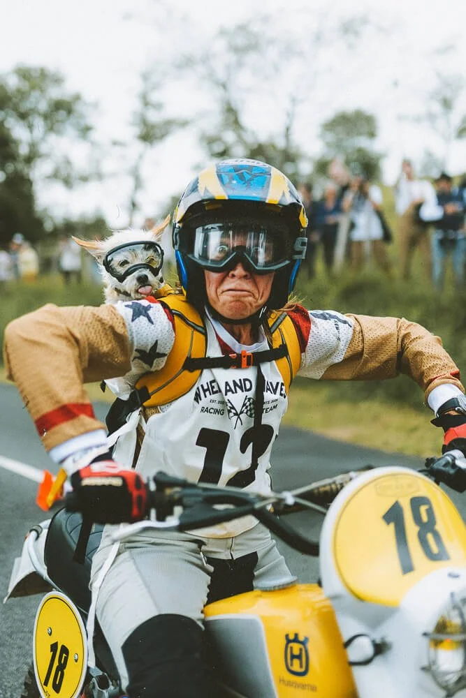 A person wearing a helmet and goggles riding a yellow and white dirt bike with the number 18 on the front. A small dog wearing goggles is perched on the rider's shoulder, sticking its tongue out. There are people in the background watching the event.