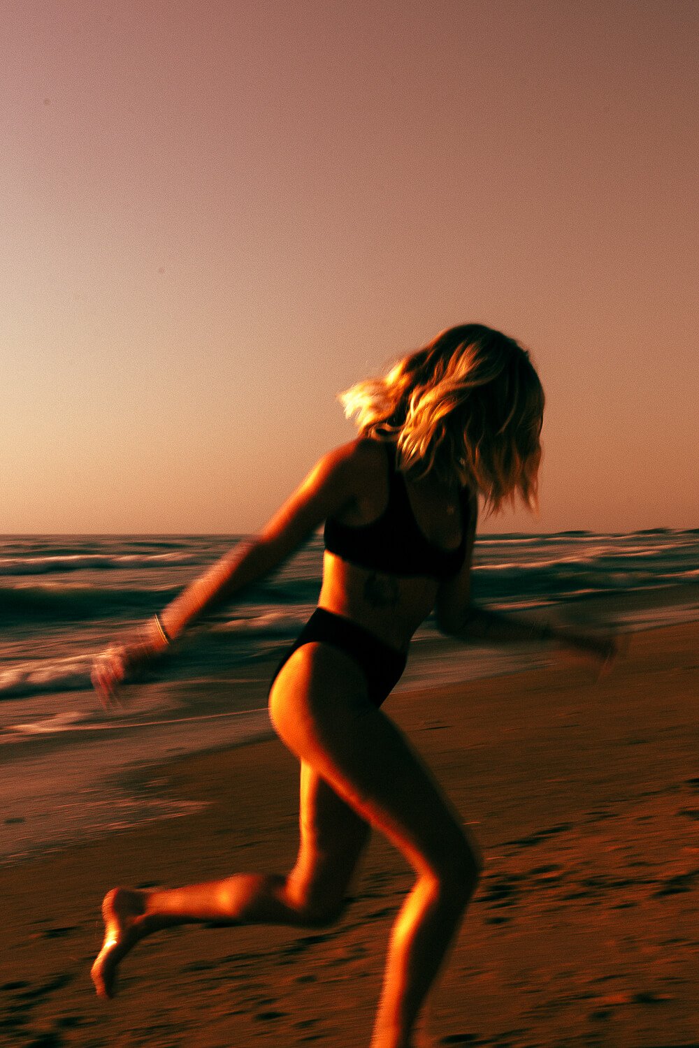 Woman in black swimsuit running on the beach at sunset.