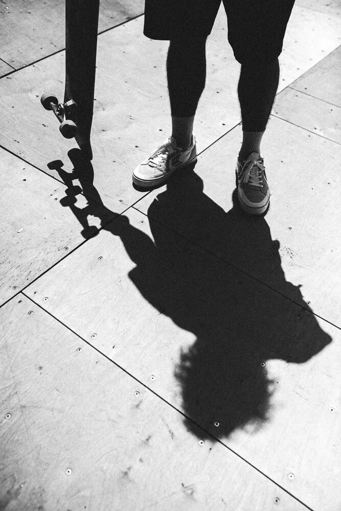 Shadow of a person with a skateboard on tiled pavement, showing their legs and shoes
