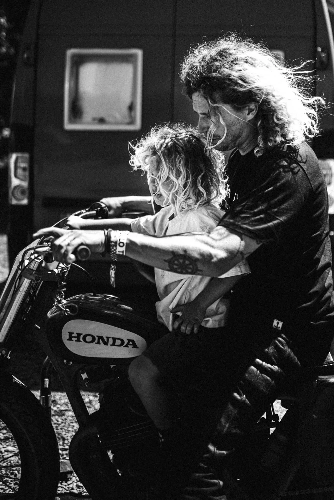 A person with long, curly hair and tattoos on their arm sitting on a Honda dirt bike, leaning forward with a child in front of them, inside a dark room.
