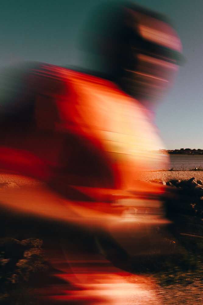 Blurry image of a person in a red shirt near a body of water during sunset.