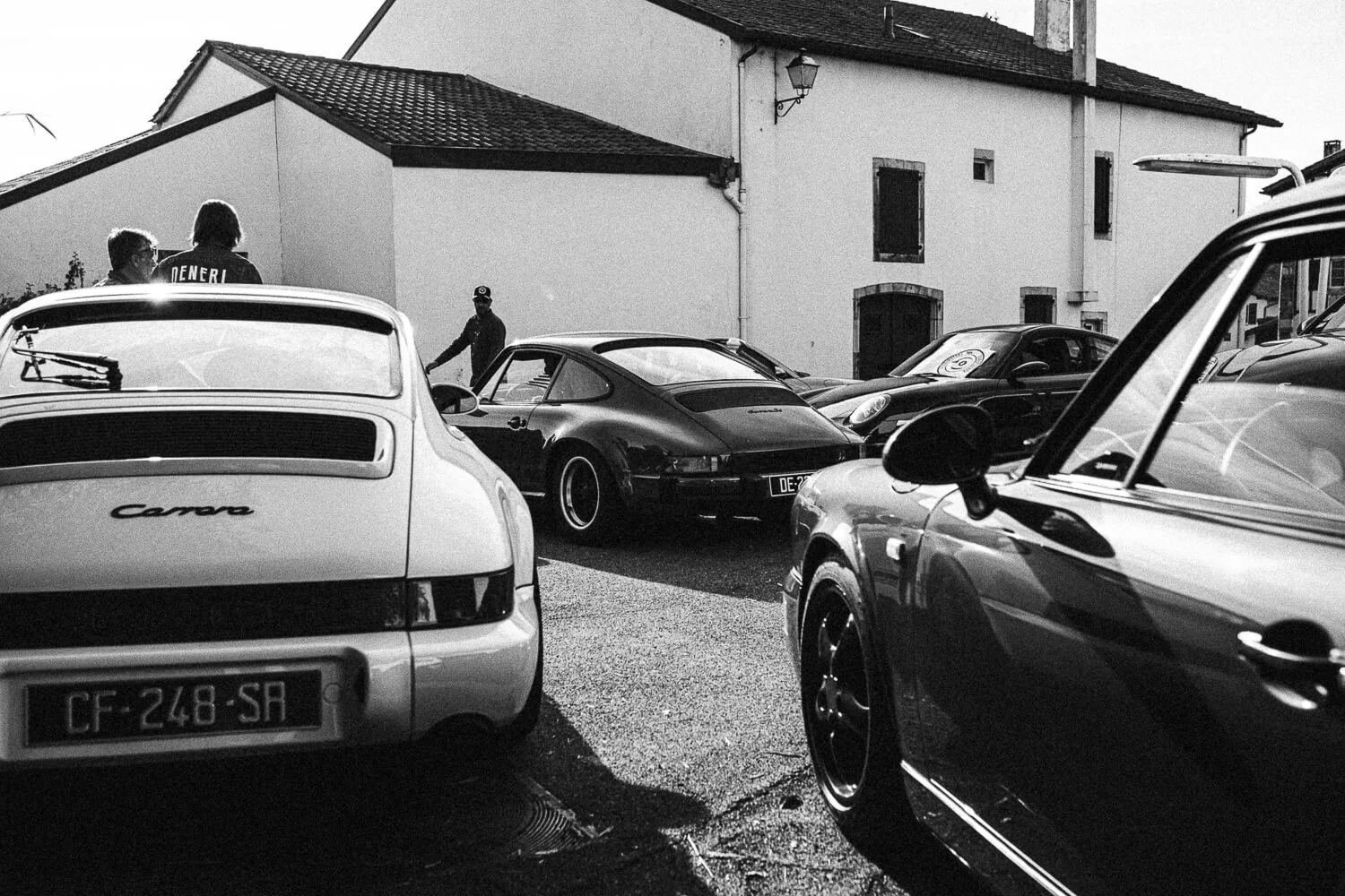 A black and white photo of a parking lot filled with classic Porsche cars, including a white Porsche Carrera with the license plate CF-248-SR and a black Porsche with the license plate DE-2. The background features a white building with a sloped roof