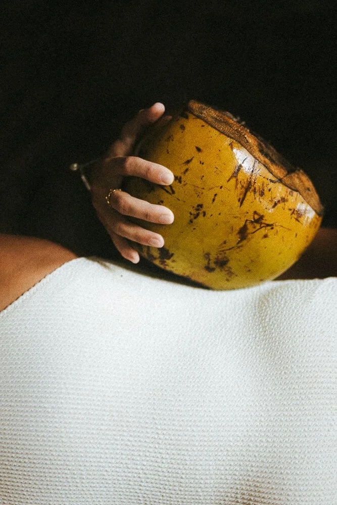 A person's hand holding a halved coconut with a yellow outer shell and brown, fibrous top, resting on a white textured cloth.