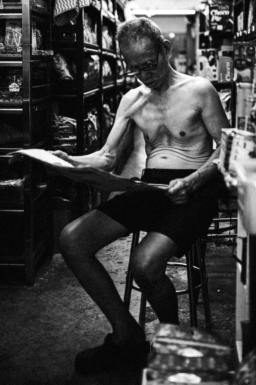 Un homme chauve et à lunettes, assis dans un magasin, sans vêtement supérieur, regardant un magazine.