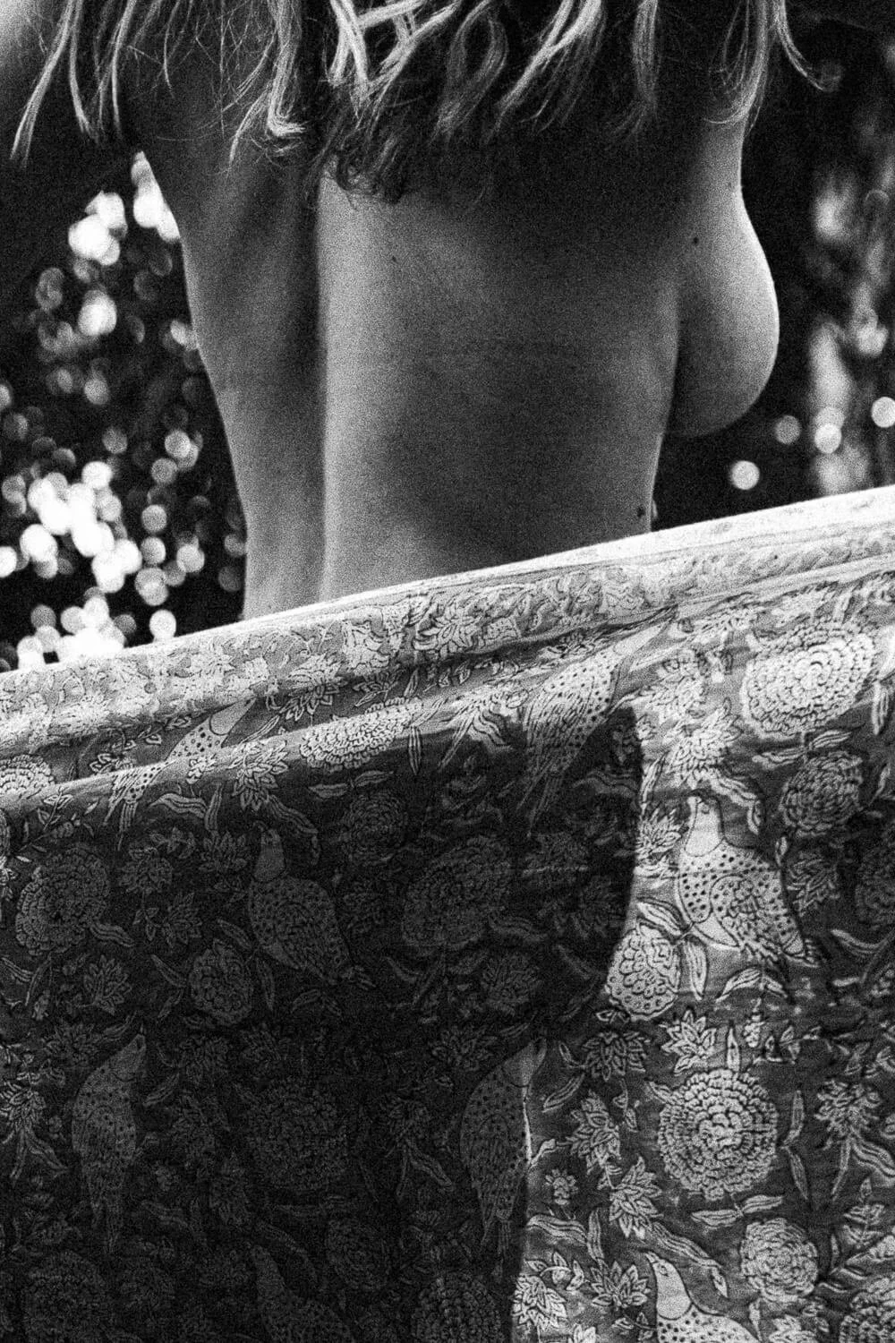 A black and white photo of a person with long hair, seen from behind, draped in a patterned fabric with floral and bird designs, with blurry lights in the background.