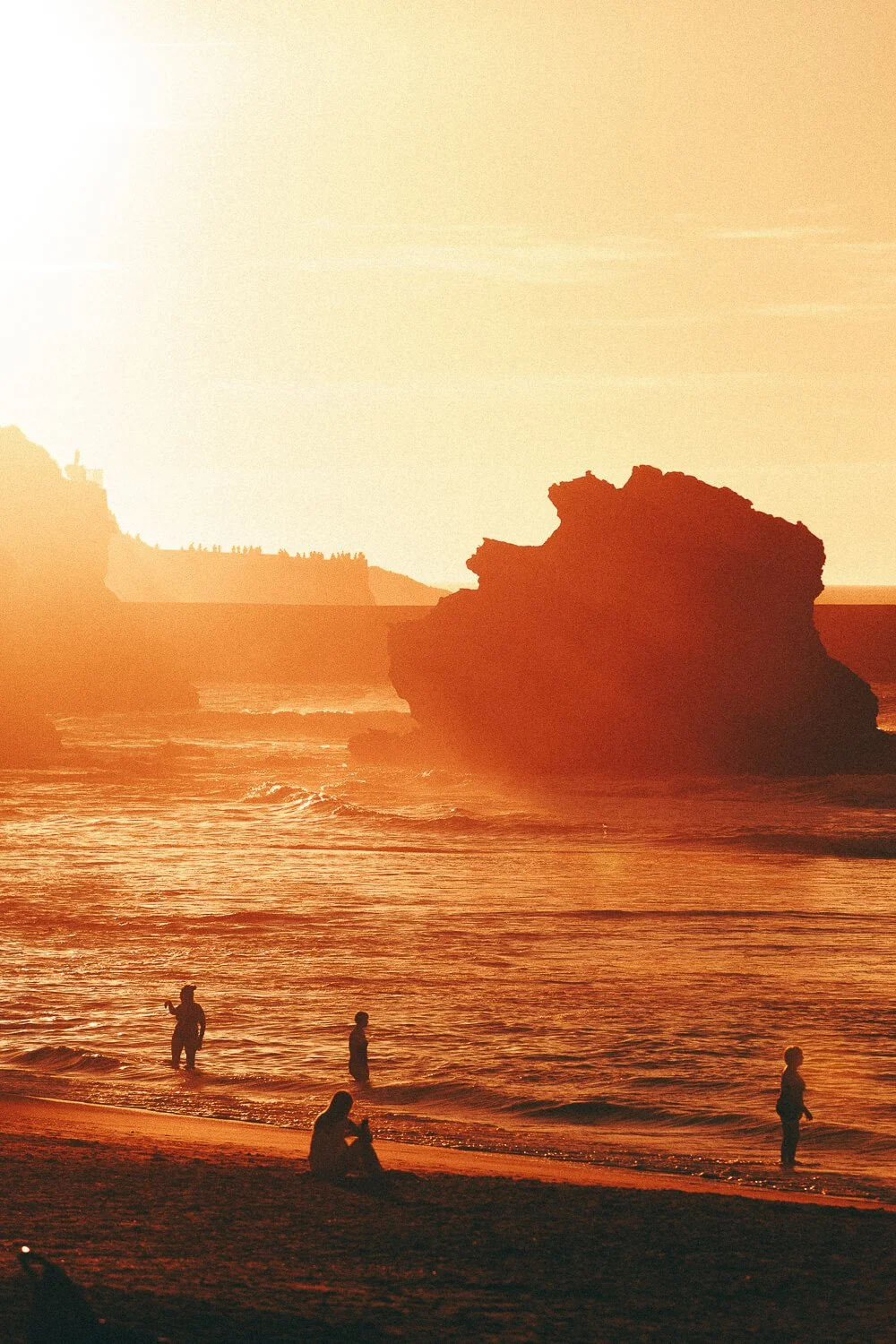 People wading and sitting on the beach during sunset, with rocky formations and cliffs in the background.