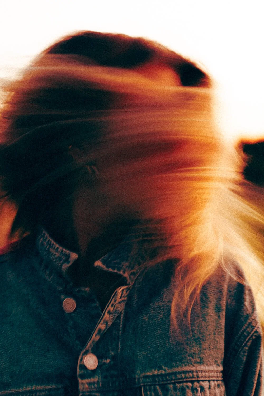 A person with long, flowing hair wearing a denim jacket, with their head turned away, creating a motion blur effect.