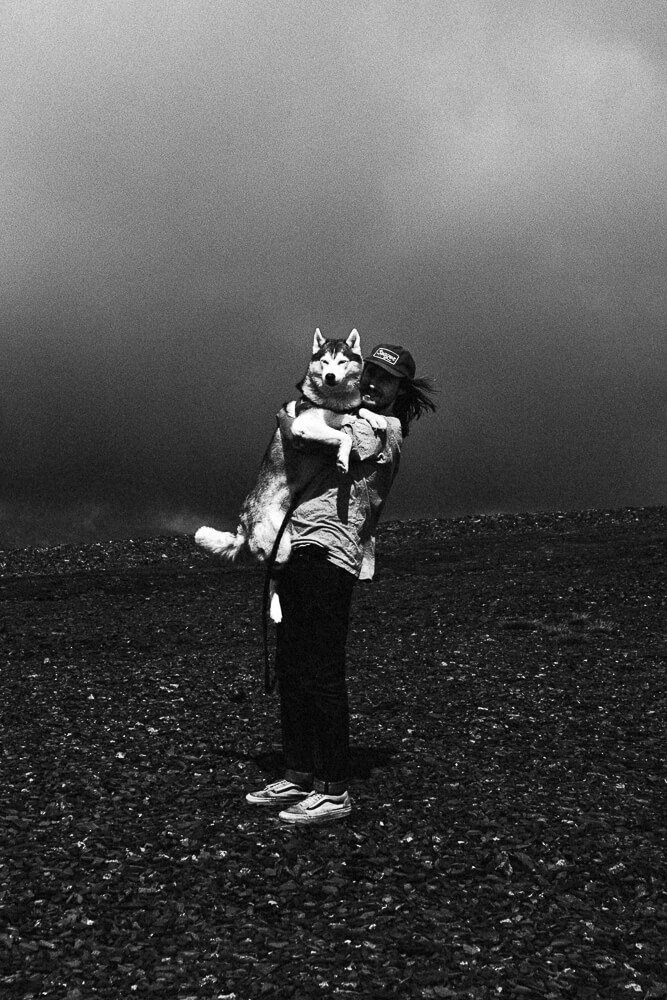 Person holding a large husky dog outdoors on a rocky surface with a cloudy sky.