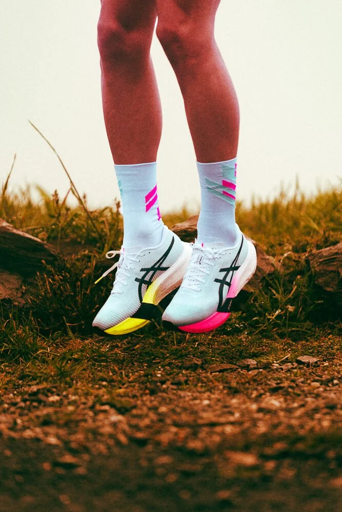 Close-up of a runner's legs wearing white running shoes with neon yellow and pink soles and white and pink socks, standing on a dirt trail with grass and rocks.