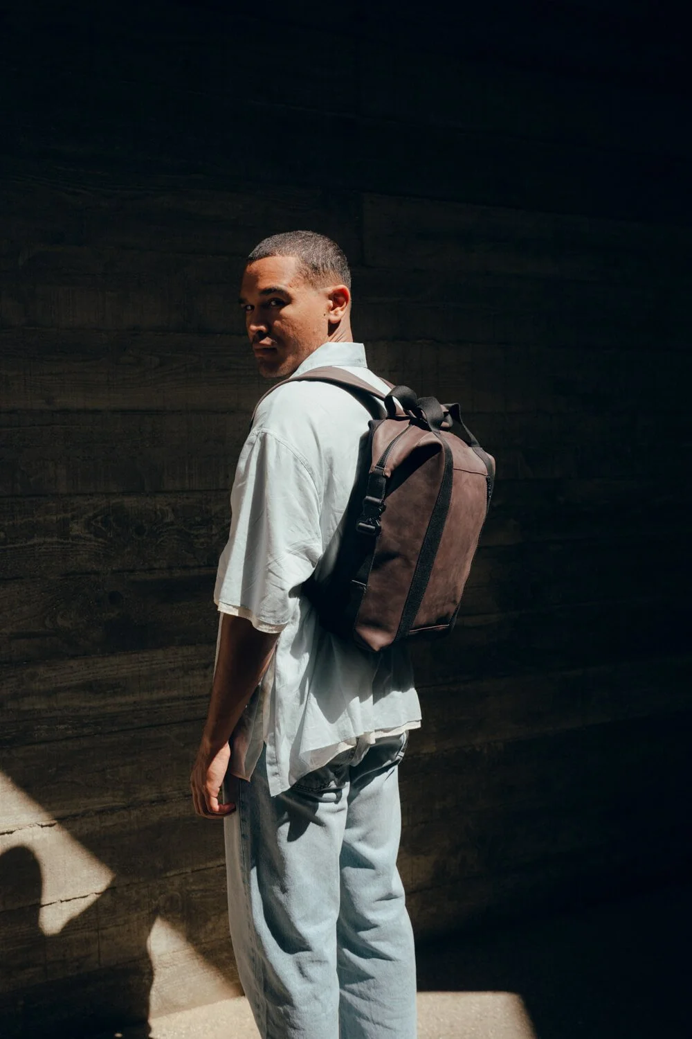 A man with short hair wearing a white shirt and light jeans, carrying a black backpack, standing against a dark textured wall with sunlight casting on him.