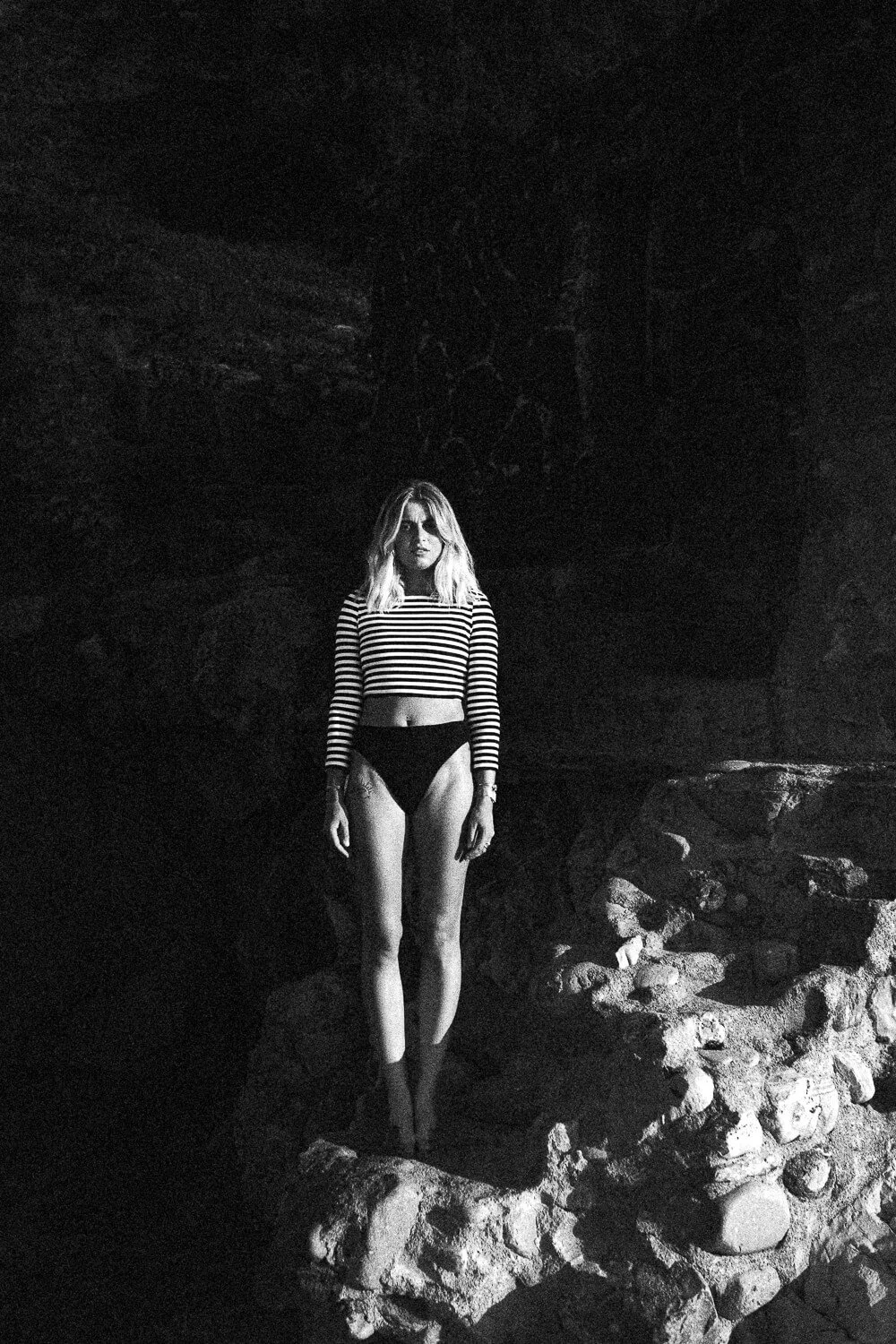 A woman with shoulder-length hair wearing a striped long-sleeved crop top and black underwear standing on rocks at night, illuminated by a light source.